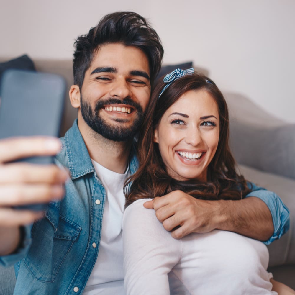 A young man and young woman sit on teh floor while the man embraces the woman from behind and they both smile while taking a cell phone photo.