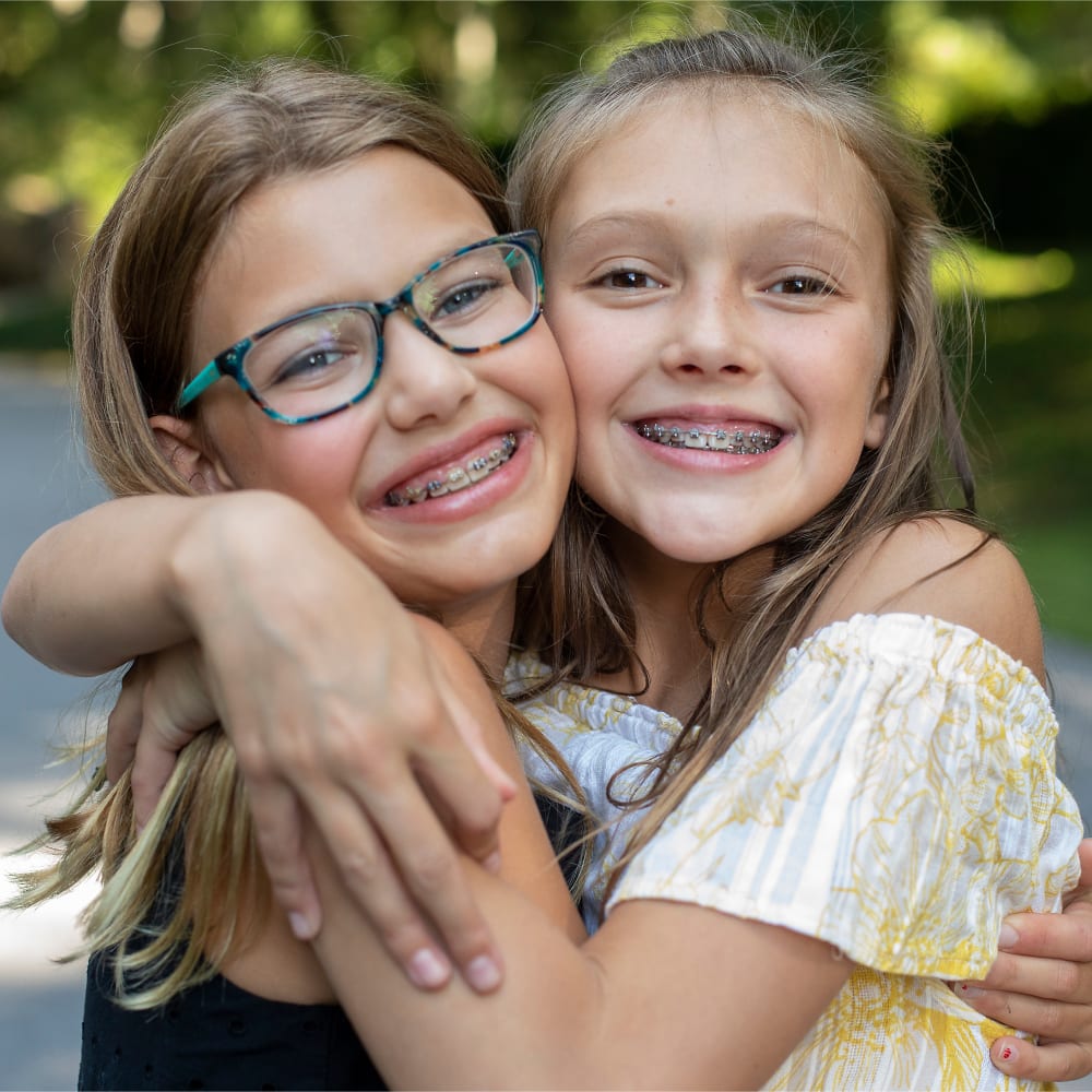A young boy wearing braces stands at a sink, smiling while a smaller boy stands behind him, just out of focus.