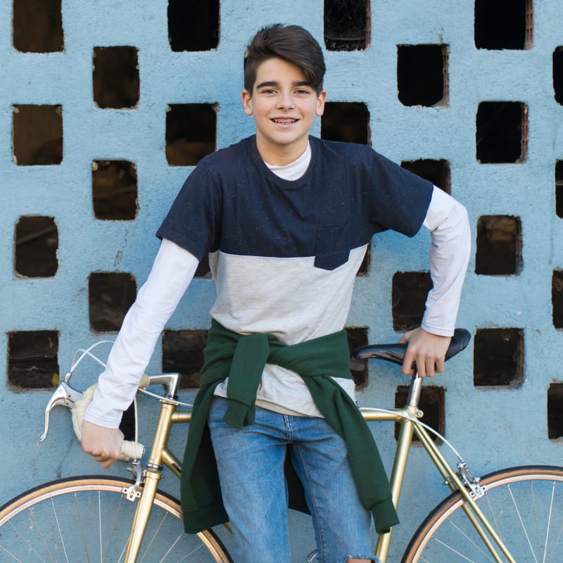 A teen boy stands in front of a blue wall, leaning on a bicycle behind him while smilng and looking at the camera.