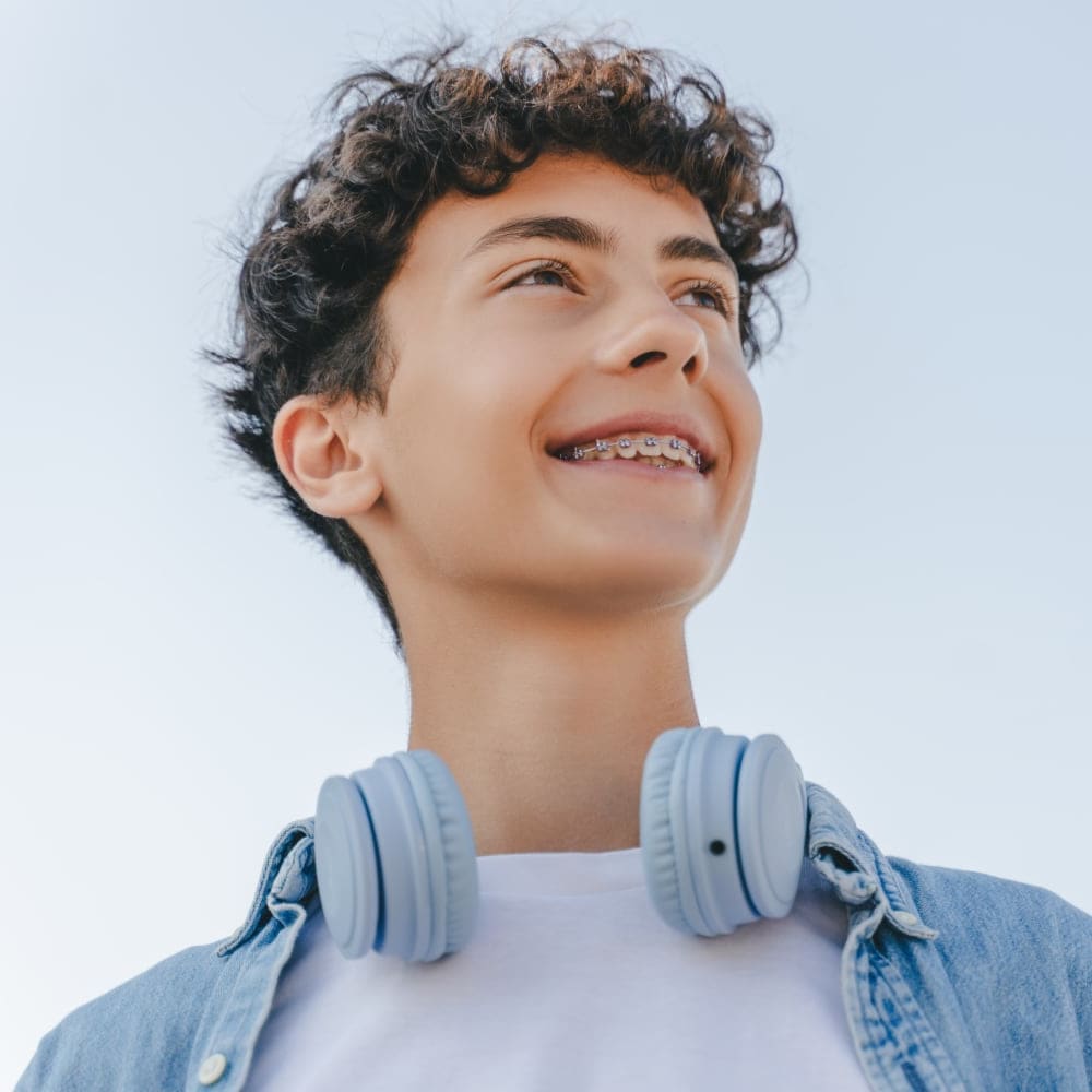 A teen boy with braces looks off to the side smiling while standing outside.