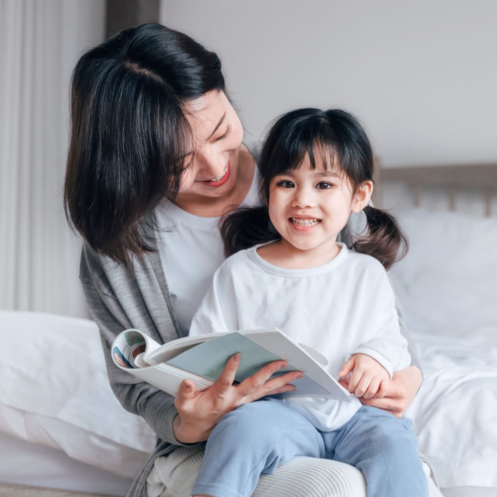 A woman sits on a bed with a small girl sitting on her lap smiling and they are looking at a book.