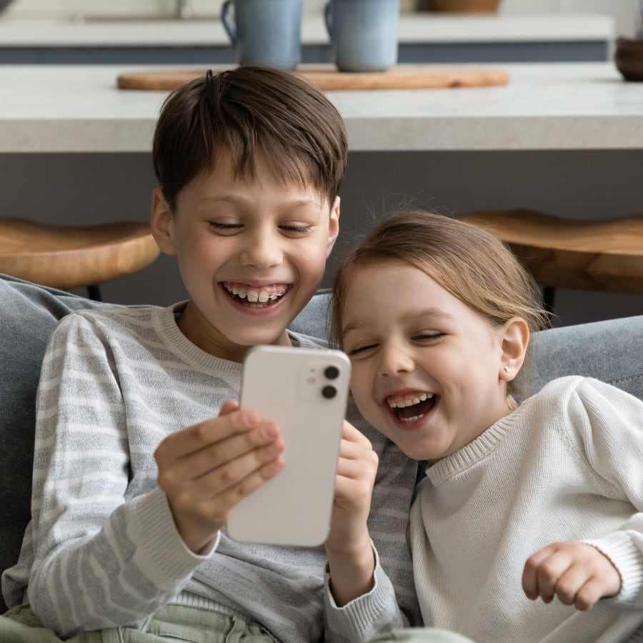 Two children sit on a sofa laughing and looking at a cellphone together,.