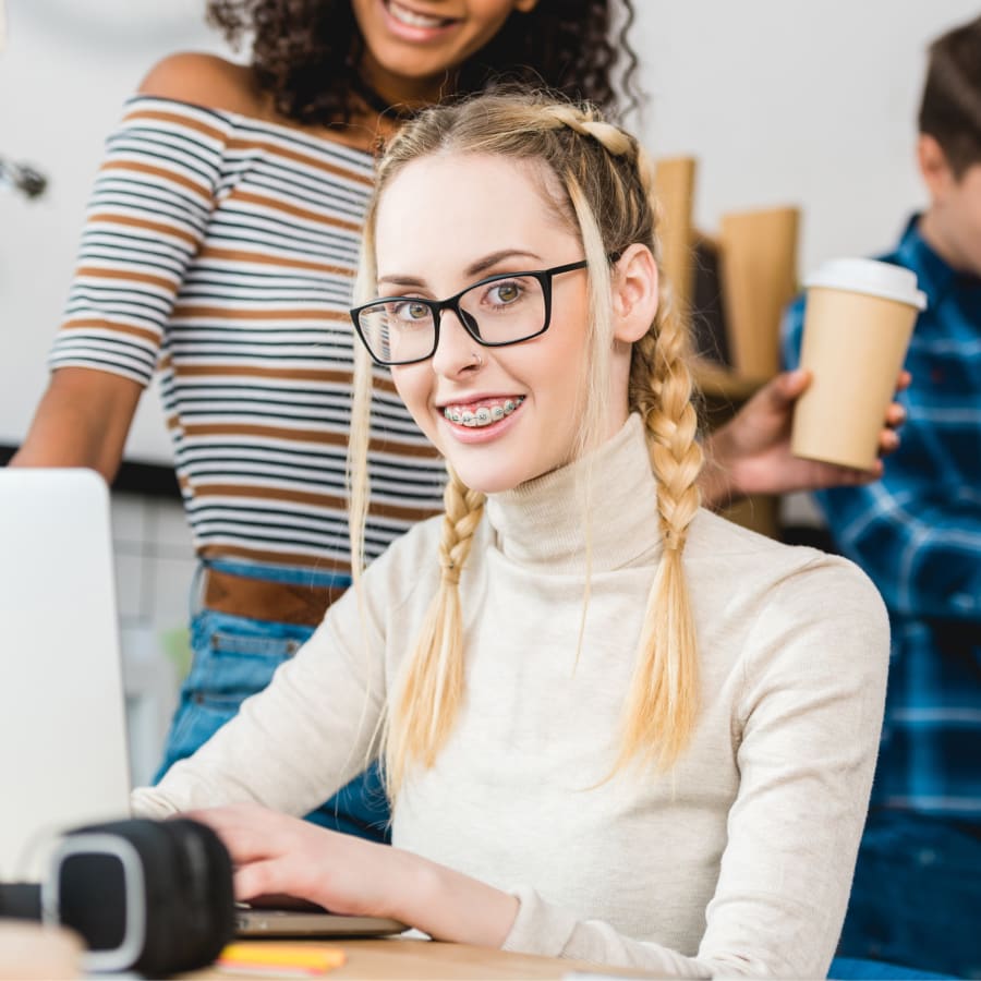A teen girl with braces typing on her laptop at school, smiling and looking at the camera