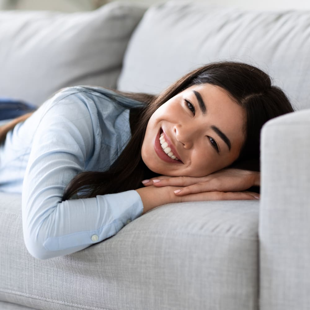 A teen girl lays on her stomach across a sofa with her hands under her face. She is looking at the camera and smiling.